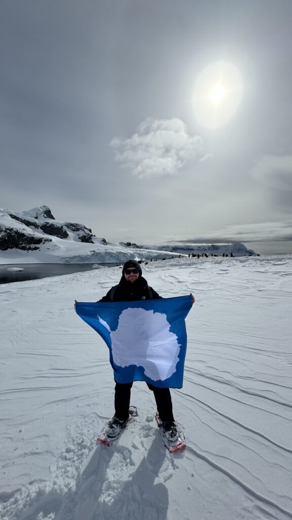 Ja z flagą Antarktydy na Cuverville Island z kolonią pingwinów w tle